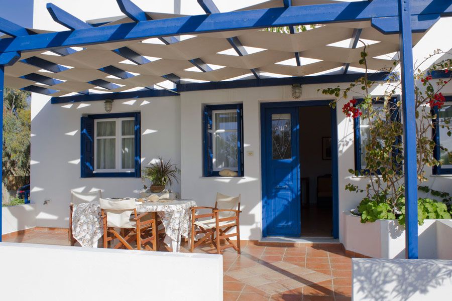 An outdoor patio space is presented under a blue trellis, featuring a circular dining table surrounded by wooden chairs. The entrance is framed by vibrant blue doors and windows, complemented by potted plants and flowering vines, with terracotta tiles adding warmth to the area.
