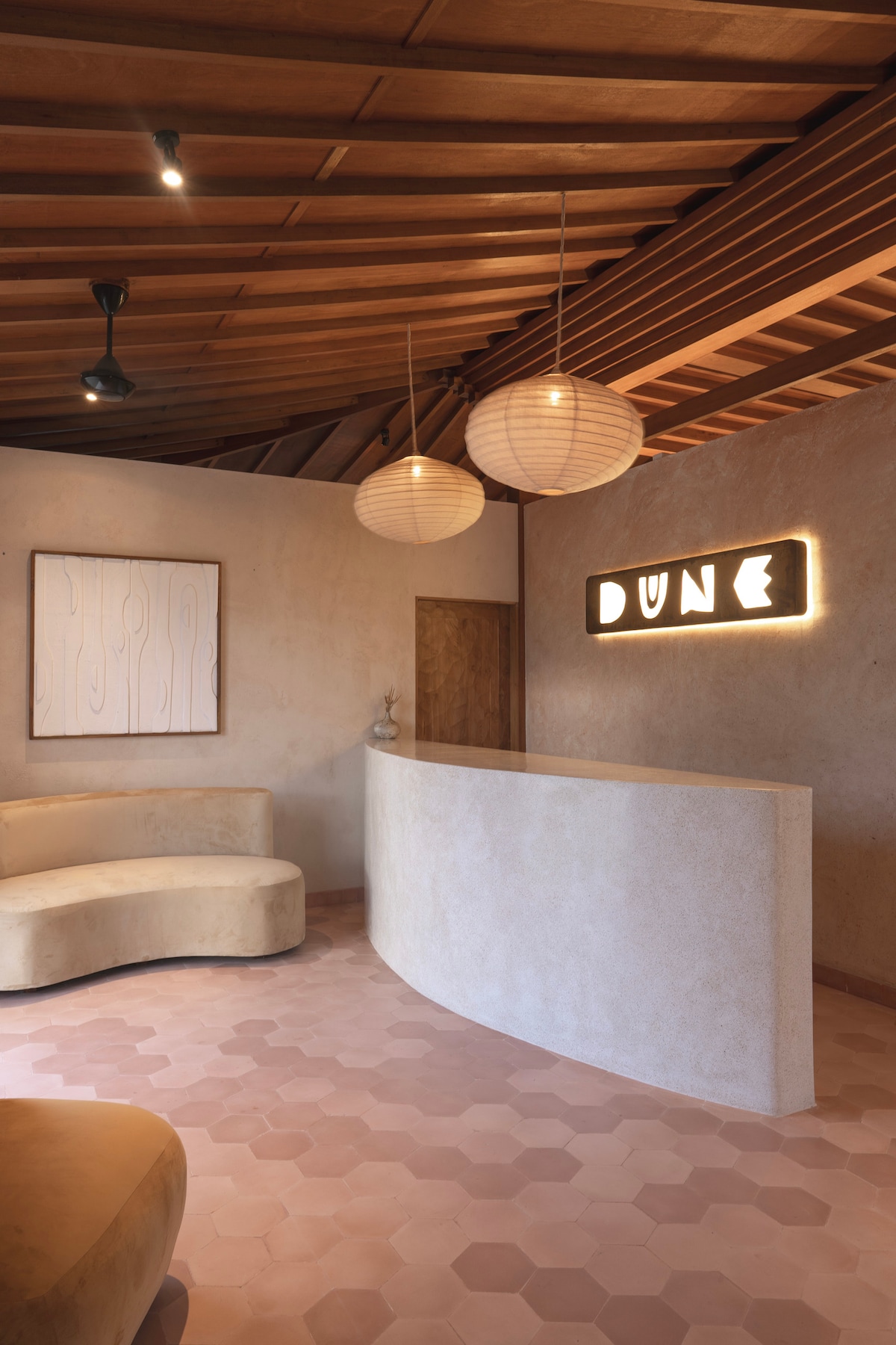 A reception area showcases a curved, textured front desk with soft, natural tones. Light wood accents and a warm ceiling create a tranquil atmosphere. Hanging pendant lights provide soft illumination, complementing a simple art piece on the wall and the stylish DUNE signage.