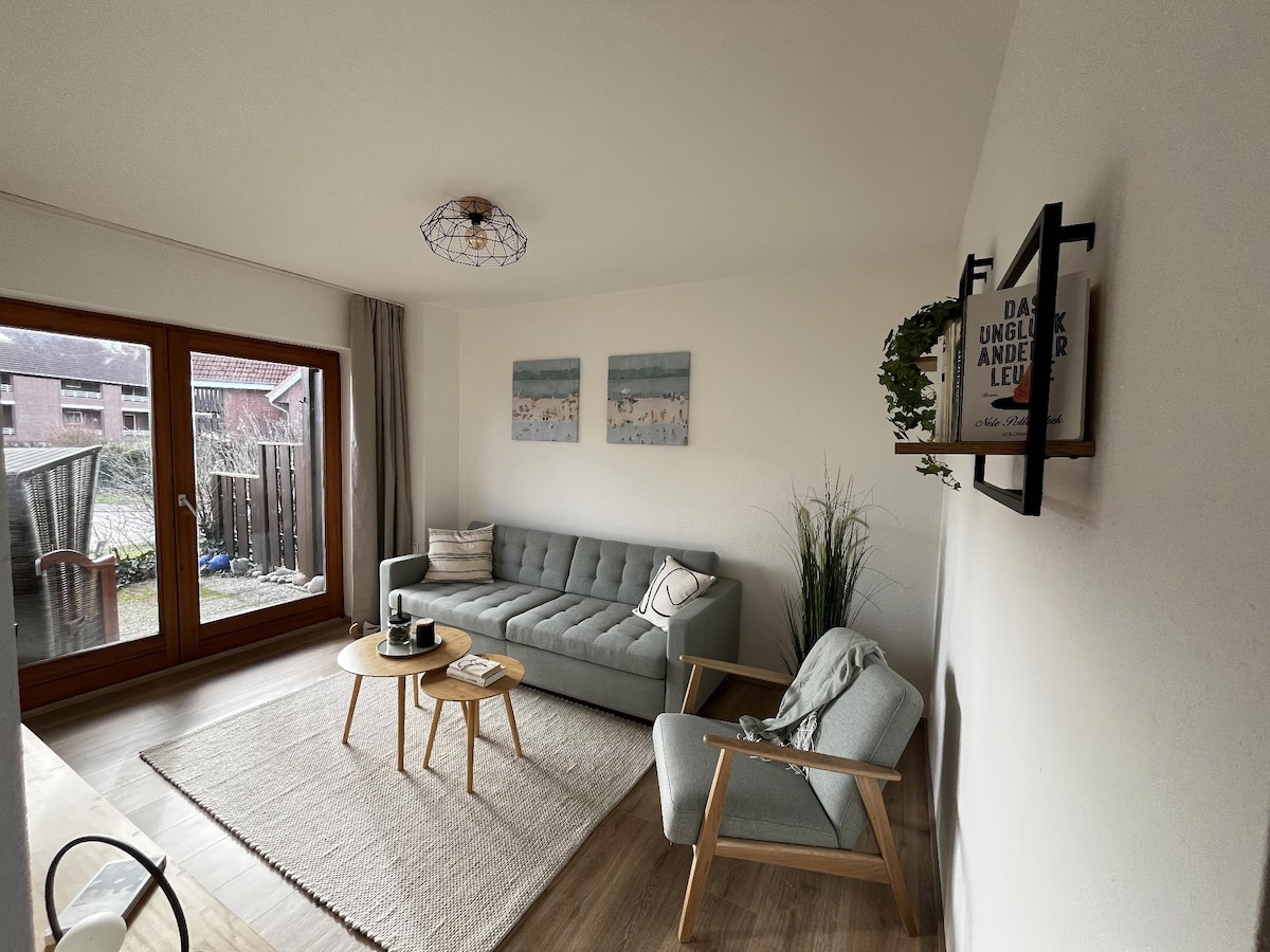 A well-lit living room features a light gray sofa and an armchair, complemented by a round coffee table. Two pieces of art adorn the wall, and large windows provide a view of the garden. A neutral-colored area rug adds warmth to the space.