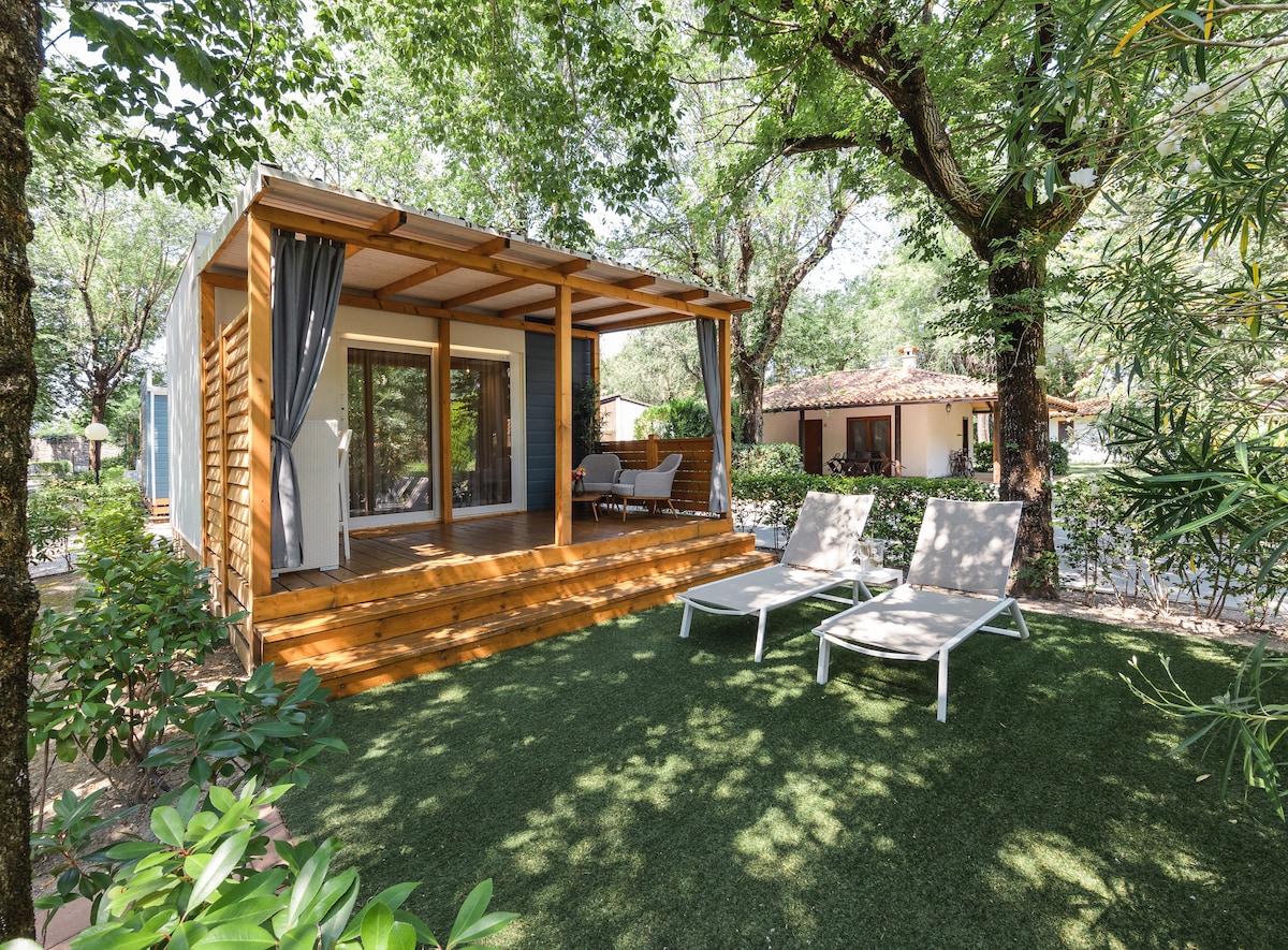 A wooden veranda is featured at the entrance, complemented by two lounging chairs. A spacious garden area with artificial grass surrounds the cottage, accented by leafy trees that provide shade, while large glass doors invite natural light into the interior.