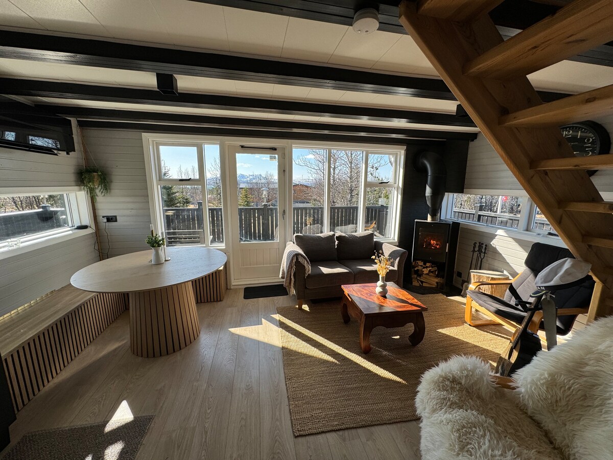 A spacious living area is characterized by natural light streaming through large windows, offering a view of the outdoors. A cozy seating arrangement includes a sofa and a chair near a modern fireplace. A round dining table is positioned adjacent, enhancing the welcoming environment.