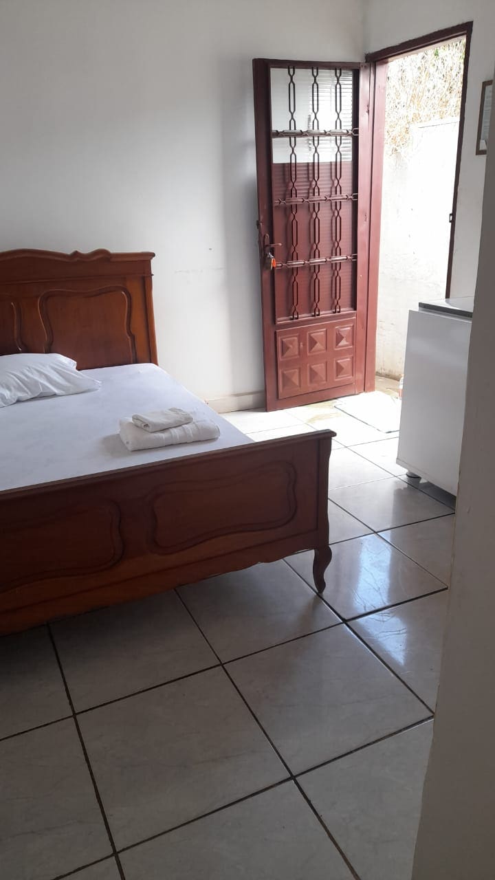 A sunlit room features a wooden bed with neatly arranged linens. A partially open door reveals a glimpse of the outdoors, while glossy tiled floors reflect the light. A small refrigerator is located nearby, adding to the room's functionality.