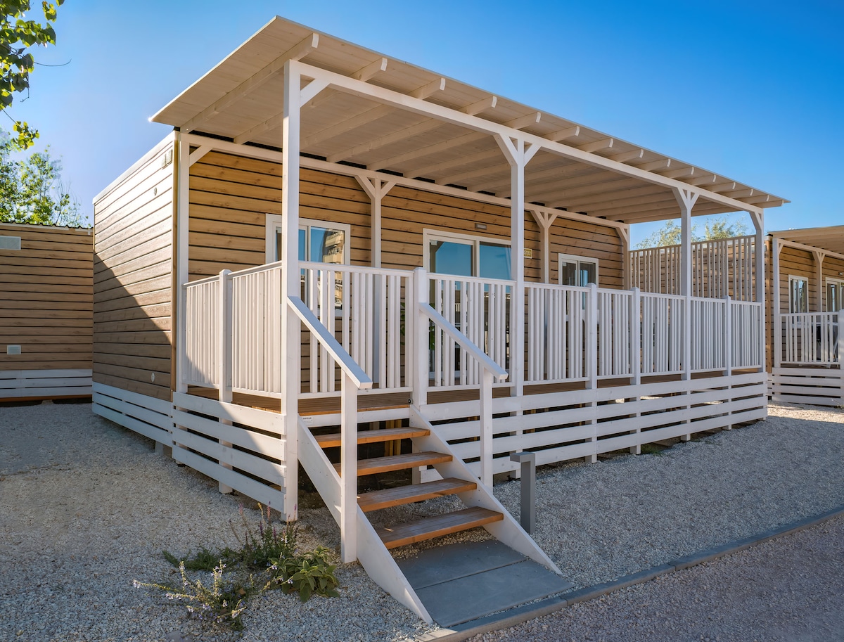 A wooden cottage features a spacious porch, surrounded by a white railing. Steps lead to the entrance, while large windows allow natural light to fill the interior. A pebble pathway adds to the outdoor landscape, providing easy access to the unit.