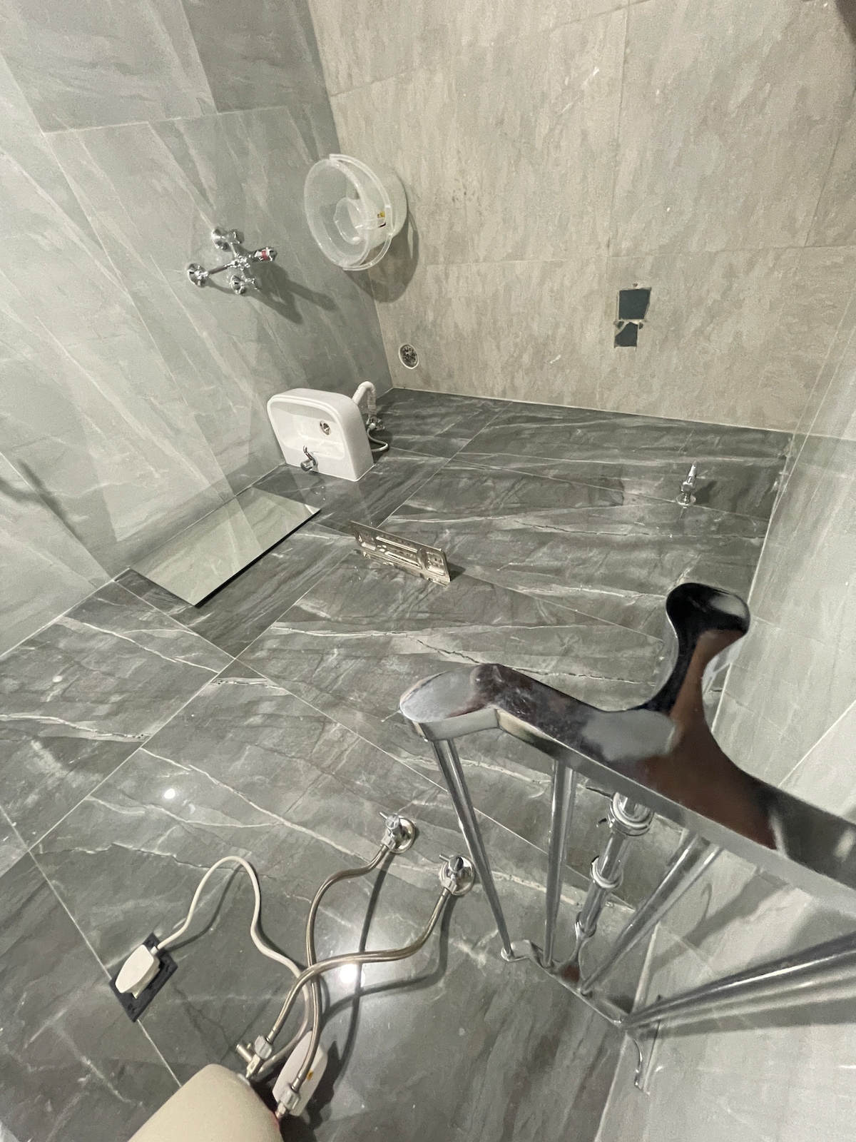 The bathroom features sleek gray marble tiling throughout the floor and walls. A toilet is positioned against one wall, and a shower area is equipped with grab bars for accessibility. Plumbing fixtures and an overhead showerhead are visible, ensuring functionality.