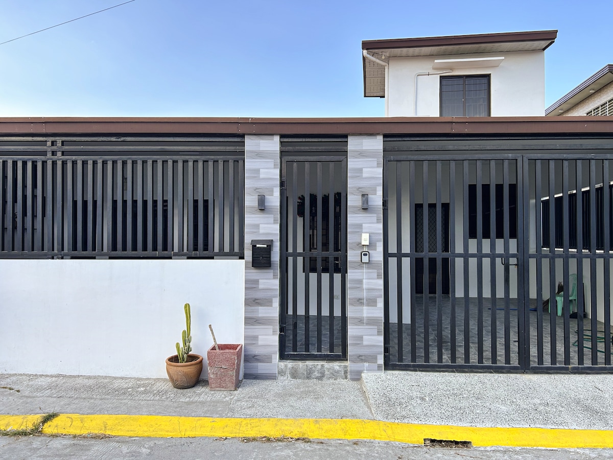 The exterior of a bungalow house is showcased, featuring a black gated entrance with vertical bars, providing a sense of security. A decorative planter with a cactus is positioned beside the entrance, complemented by a light-colored façade and concrete detailing. A clear sky frames the home.