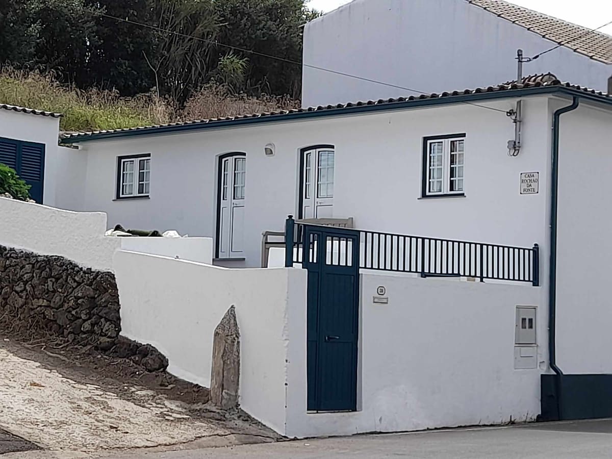 A charming white house with a blue door and window shutters is set against a natural landscape. A simple railing encloses the small patio area, while the well-maintained exterior features a combination of stone and stucco elements.