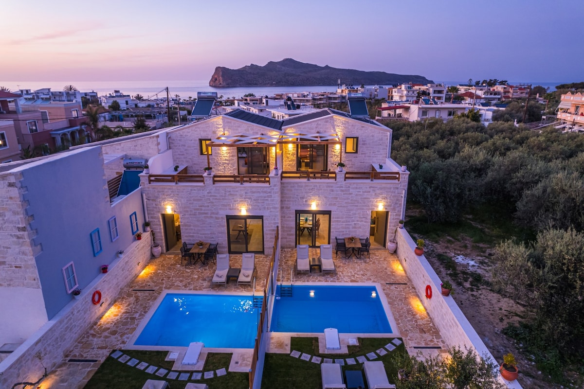 The image captures a view of two villas, each with its own private pool, set against a backdrop of the sea and a distant island. Soft evening lighting highlights the stone facades and outdoor lounging areas, while a landscaped garden area adds to the tranquility of the scene.