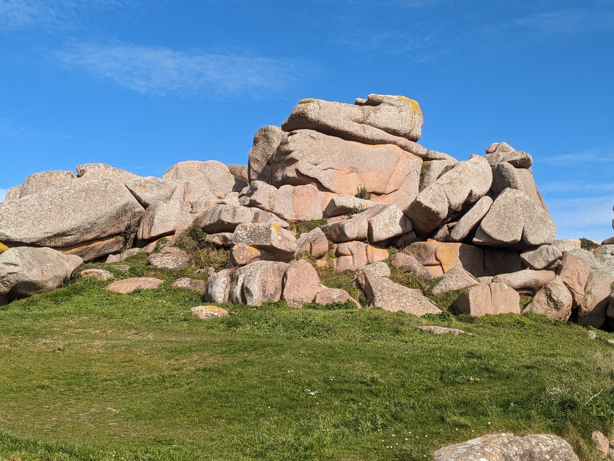 A natural landscape features a cluster of large granite rocks arranged on a grassy area. The rocks vary in size and exhibit shades of pink and gray, contrasting with the vibrant blue sky above. The scene evokes a sense of tranquility and connection to nature.