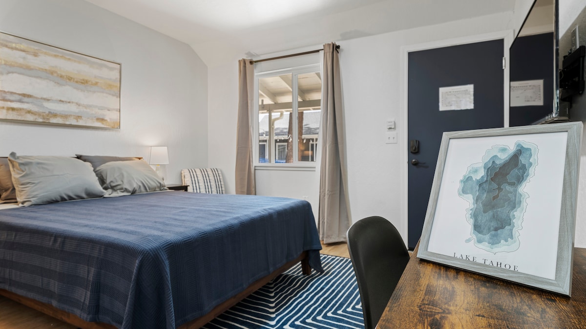 A contemporary room features a full-sized bed with a textured blue blanket and gray pillows. Natural light fills the space from a nearby window adorned with beige curtains. A desk with a decorative map of Lake Tahoe and a sleek black door is visible.