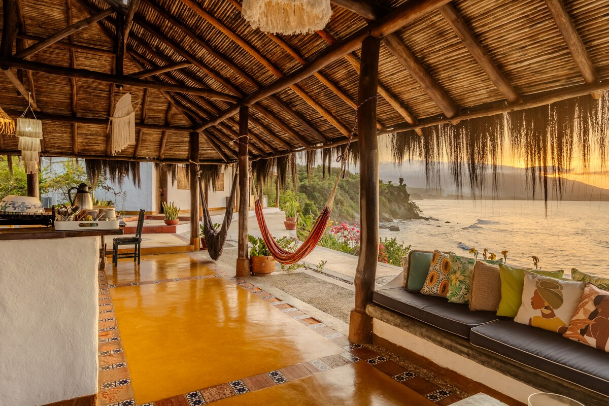 An open-air living space features a thatched roof and wooden beams, complemented by a cozy seating area adorned with colorful cushions. Hammocks are suspended nearby, and the ocean is visible beyond the lush greenery, providing a serene backdrop for relaxation.