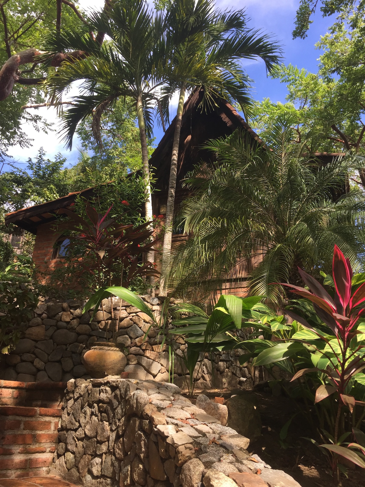 A charming Balinese casita is nestled among vibrant foliage, featuring a thatched roof and wooden exterior. Lush tropical plants surround the structure, and stone pathways lead to the entrance, creating a serene and inviting atmosphere in the jungle.