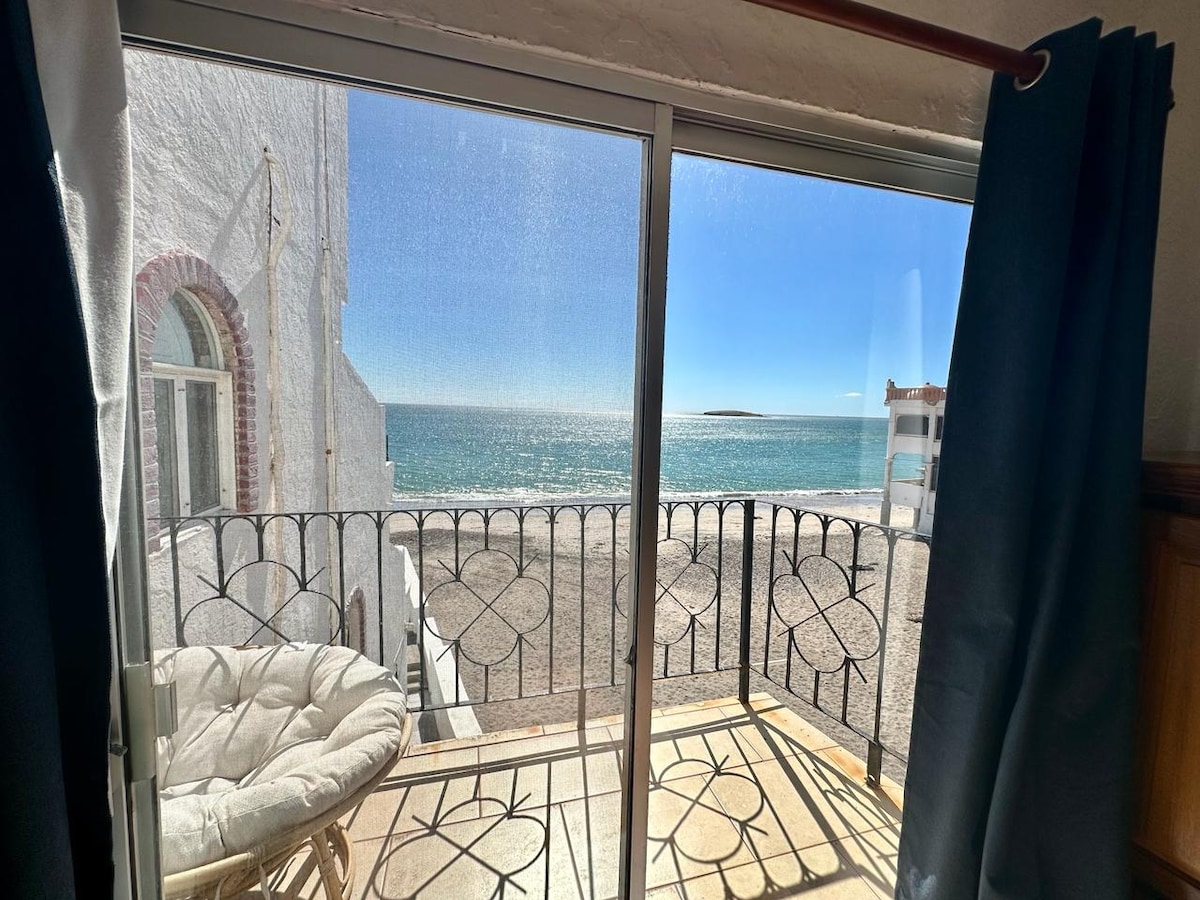 A sliding glass door opens to a private balcony, revealing expansive ocean views. Decorative ironwork frames the view, while natural light filters through sheer curtains. The serene shoreline and clear blue water enhance the inviting atmosphere of the space.