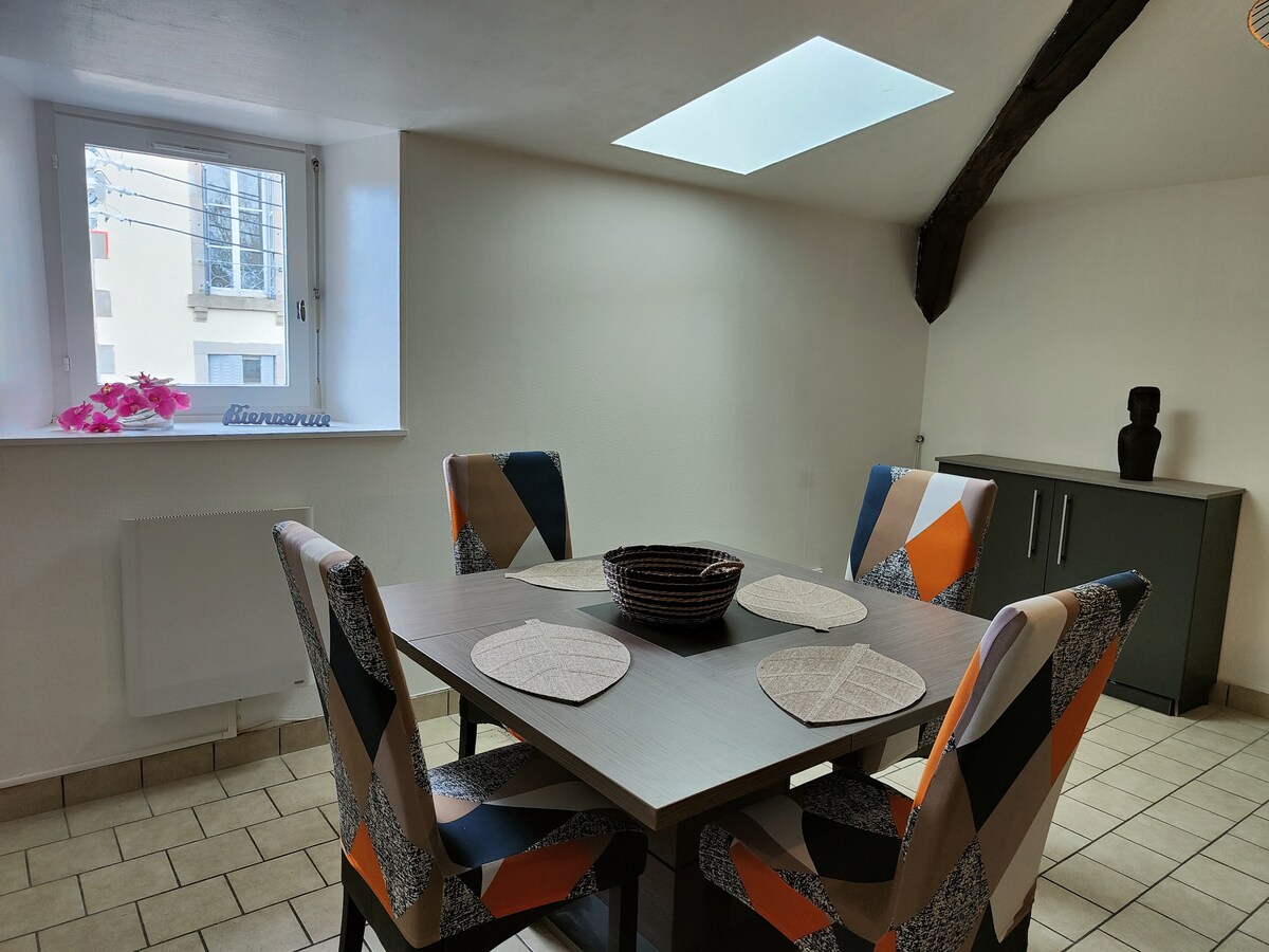 A dining area is showcased with a rectangular table surrounded by four chairs featuring colorful geometric patterns. Natural light enters through a skylight and a nearby window, illuminating the light-colored walls and tiled floor.