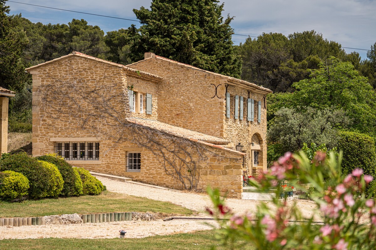 A charming stone mas is presented with classic architectural features, including light-colored shutters and a tiled roof. Neatly landscaped gardens with lush greenery and flowering plants surround the exterior, complemented by a serene pathway leading to the entrance.