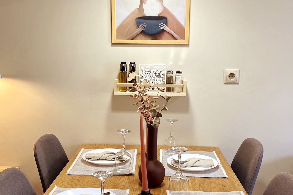 A dining area features a wooden table set with plates and wine glasses. A minimalistic vase holds decorative branches at the center. Above the table, a wall-mounted shelf displays cooking oils and spices, complemented by a framed artwork of a person holding a bowl.