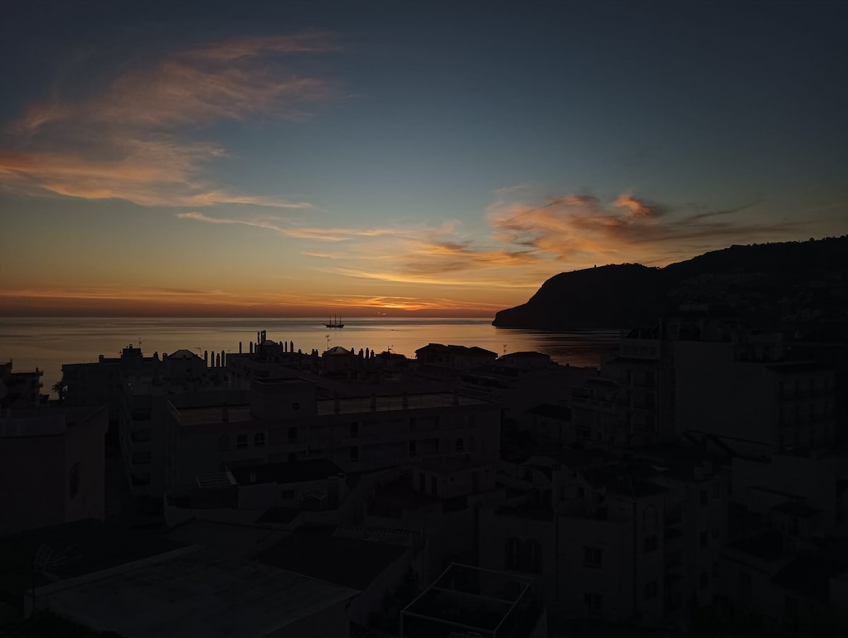 An evening view captures a serene sunset over the water, with soft hues of orange and blue illuminating the sky. Silhouettes of buildings and distant hills frame the coastline, complementing the tranquil ambiance of the scene.