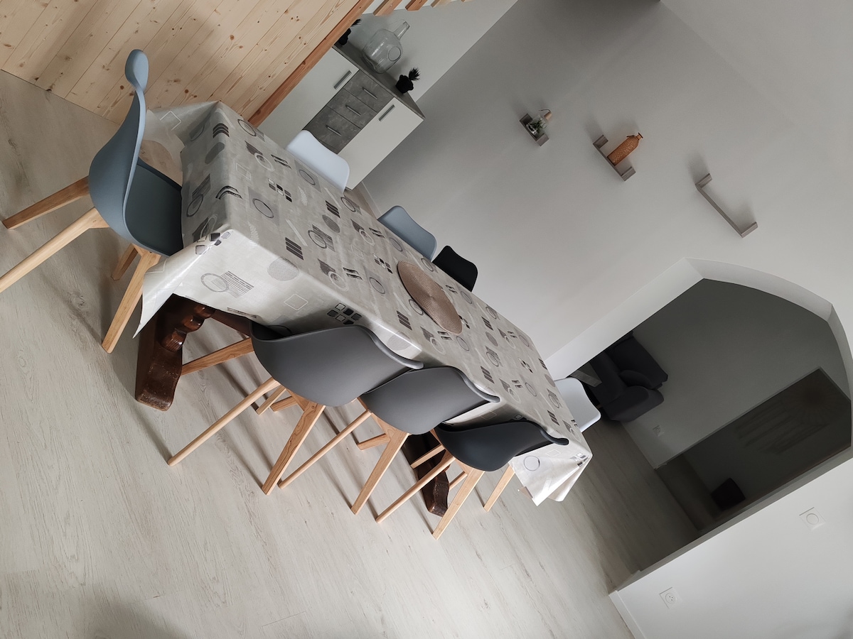 A dining area is presented with a long table covered in a patterned cloth. Several chairs, in varying shades of gray and white, surround the table. Light-colored wood flooring complements the bright, neutral walls and the open layout of the space.