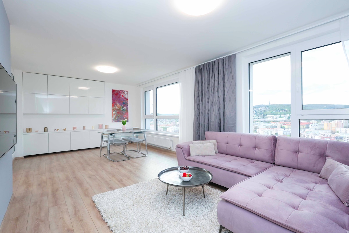 A contemporary living area features large windows that allow ample natural light to fill the space. A plush purple sectional sofa invites relaxation, while a round coffee table centered on a light-colored rug complements the modern dining area in the background.