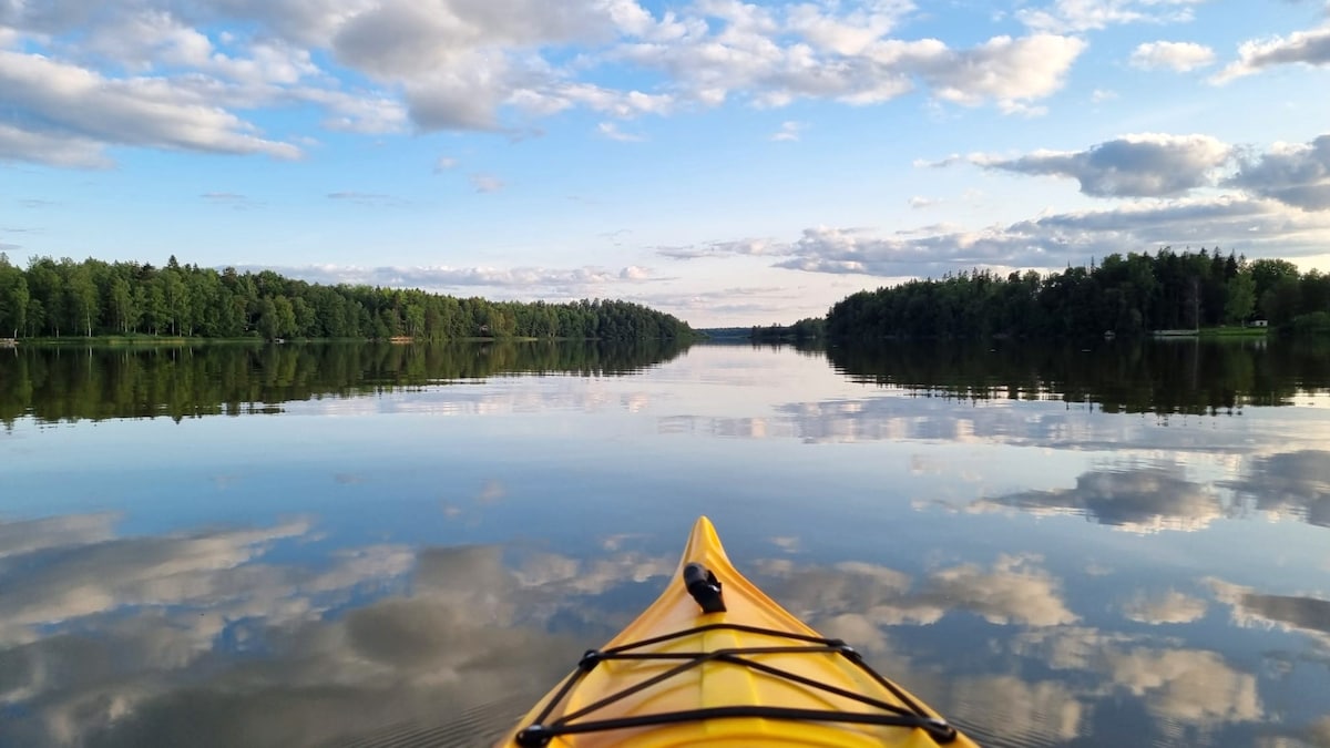 A vibrant yellow kayak is positioned at the forefront, gliding smoothly across a calm lake. Its surface reflects the surrounding green forests and soft clouds, creating a serene and picturesque scenery. The clear sky above enhances the tranquil setting, inviting exploration of the peaceful waters.