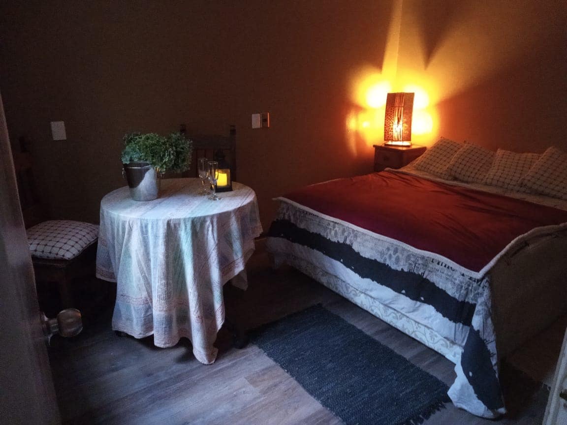 A warm bedroom environment is presented, featuring a spacious bed with a textured blanket. A small round table with a tablecloth is positioned beside it, complemented by a potted plant and softly glowing lantern. The walls are painted in earth tones, enhancing the tranquil setting.