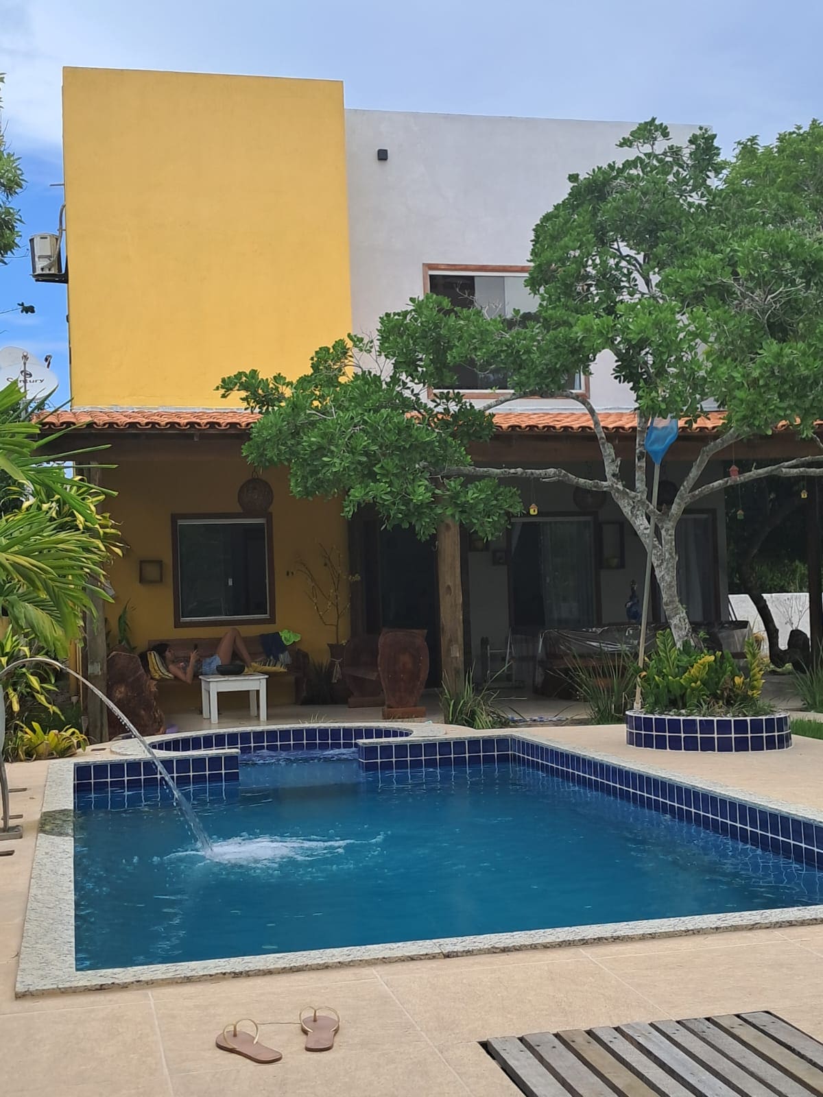 A refreshing swimming pool is framed by vibrant greenery and a spacious patio area. The two-story house features large windows and a covered terrace with wooden beams, blending indoor and outdoor living. Cozy seating areas are visible, inviting relaxation under shaded spots.