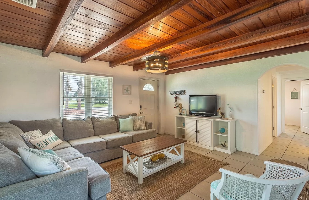 A cozy living room features exposed wooden beams overhead and soft natural light from a nearby window. A spacious gray sectional sofa is paired with a wooden coffee table and decorative elements. A TV stand holds a flat-screen TV, complementing the welcoming atmosphere.