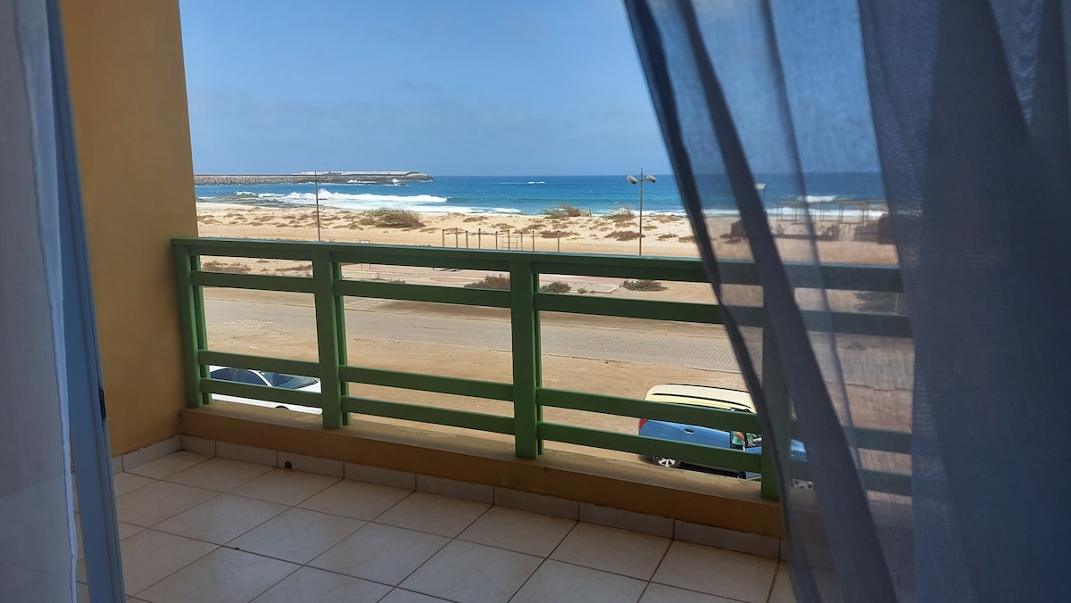 A balcony offers an unobstructed view of the ocean, with gentle waves lapping at the shore. Light sheer curtains drape the entrance, softening the natural light that filters into the space. The surrounding area features sandy beaches and distant coastal structures.