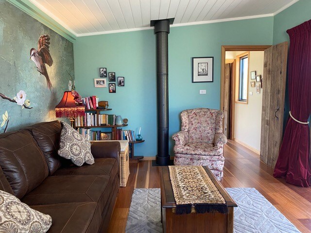 A sitting area features a brown leather couch with decorative cushions, a floral armchair, and a wooden coffee table. A wood heater is positioned against a teal wall adorned with bird-themed art. Bookshelves filled with books line the wall, enhancing the cozy ambiance.