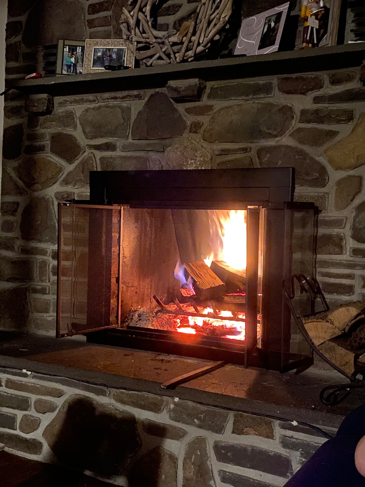 A cozy fireplace is framed by a stone mantel, with logs burning gently inside. The warm glow of the flames contrasts with the textured stone, creating a calming focal point. Above, photographs and decorative items rest on the mantel, adding to the cabin's inviting atmosphere.