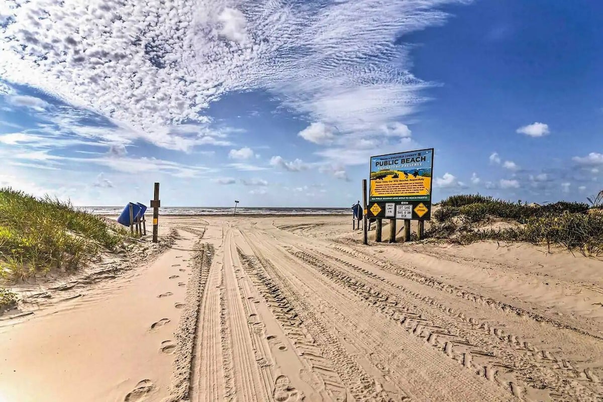 A sandy path leads to a public beach, flanked by grass and signage indicating beach rules. The sky is dotted with clouds, creating a bright and open atmosphere with ocean views in the distance. Footprints in the sand hint at recent visitors.