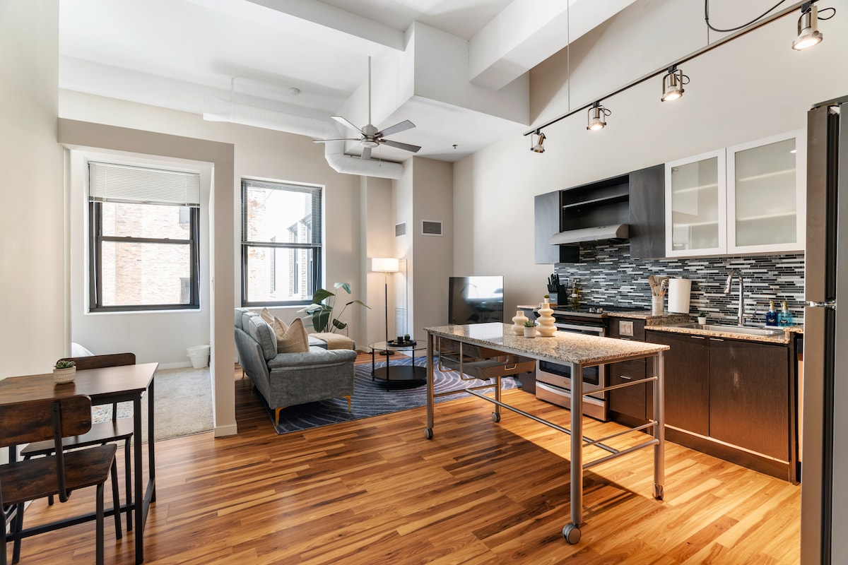 An open-concept living area showcases sleek wooden flooring and modern furnishings. A cozy sofa is positioned near a large window, allowing natural light to fill the space. The kitchen features stylish cabinetry, an island, and high-quality appliances, creating an inviting atmosphere for cooking and entertaining.