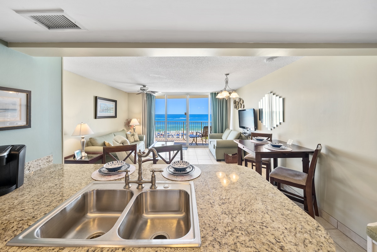 A modern kitchen with a granite countertop features a double sink and is oriented toward a living area with panoramic Gulf views. The space includes a cozy seating area and a dining table set for two. Large windows allow abundant natural light to fill the room.