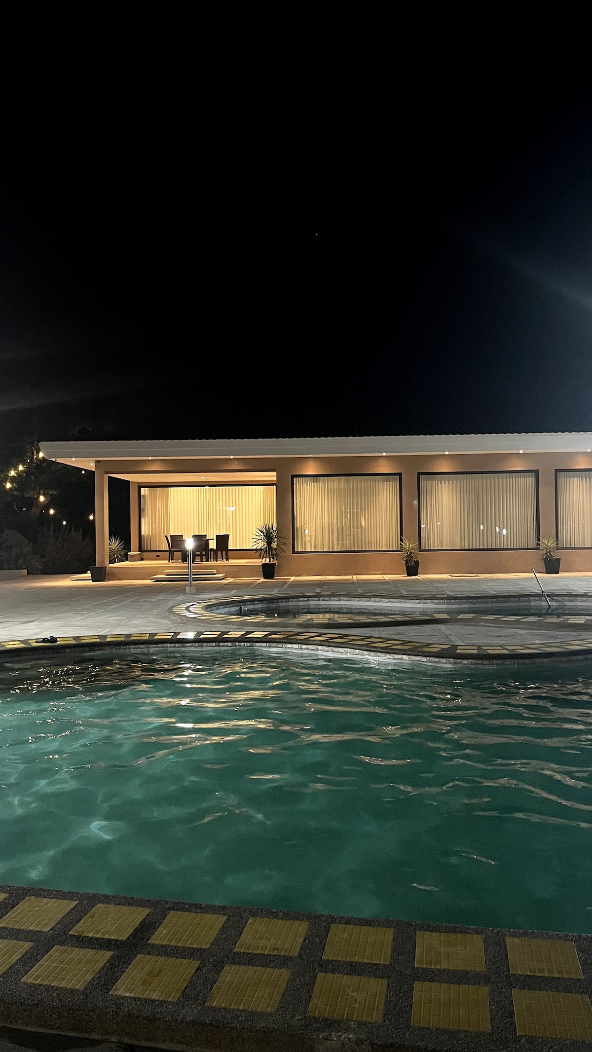 A nighttime view of a modern structure featuring large windows and patio lights. A clear pool reflects the gentle illumination, and potted plants are placed strategically along the building's exterior, enhancing the serene environment.