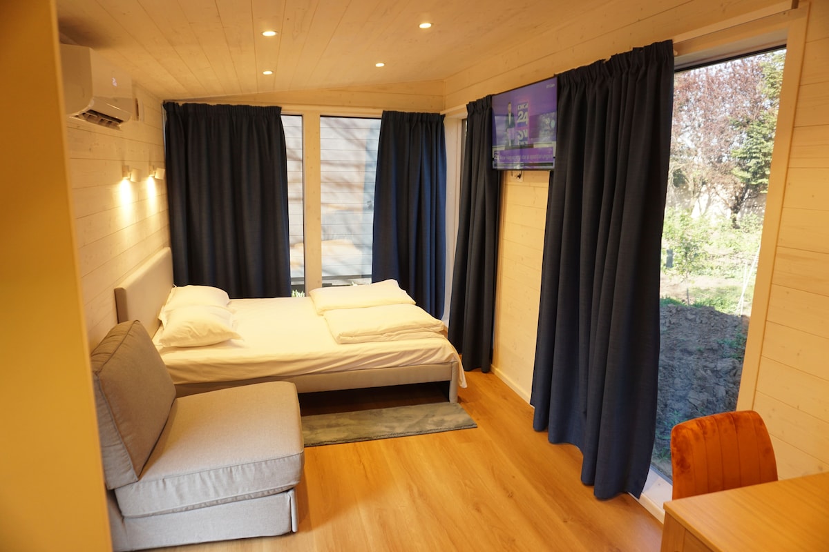 A bright interior features a spacious bed situated beside a cozy sofa. Floor-to-ceiling windows are dressed with dark curtains, allowing natural light to filter in. A wall-mounted Smart TV is positioned across from the seating area, contributing to a modern living space.