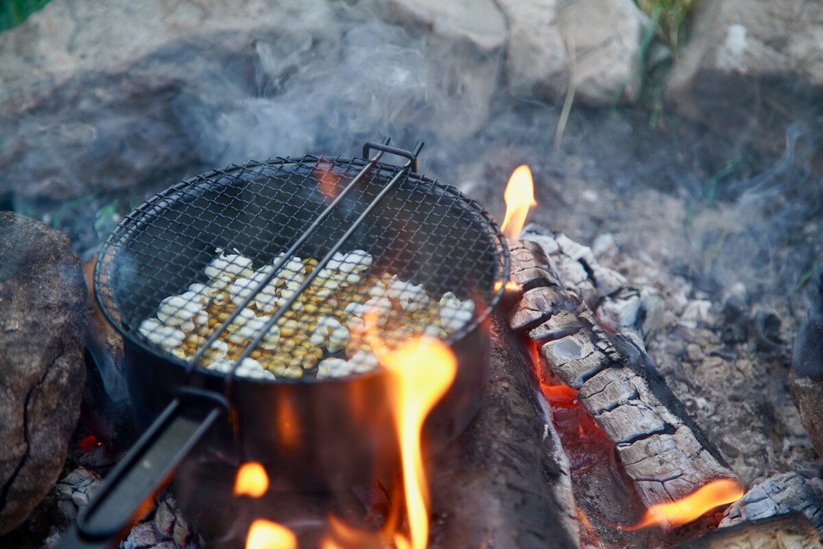 A grill basket sits over an open fire, displaying an assortment of food being cooked. The flames create a warm glow, while smoke rises gently from the campfire. Stones are arranged around the fire, adding to the rustic outdoor setting.