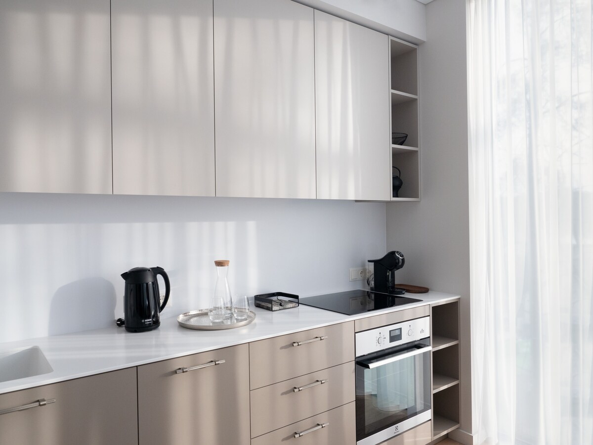 A modern kitchen space is presented, featuring sleek cabinetry and a countertop. Kitchen appliances include a kettle, coffee machine, and oven with an induction cooktop. Windows with sheer curtains allow natural light to flow in, enhancing the bright and airy feel.