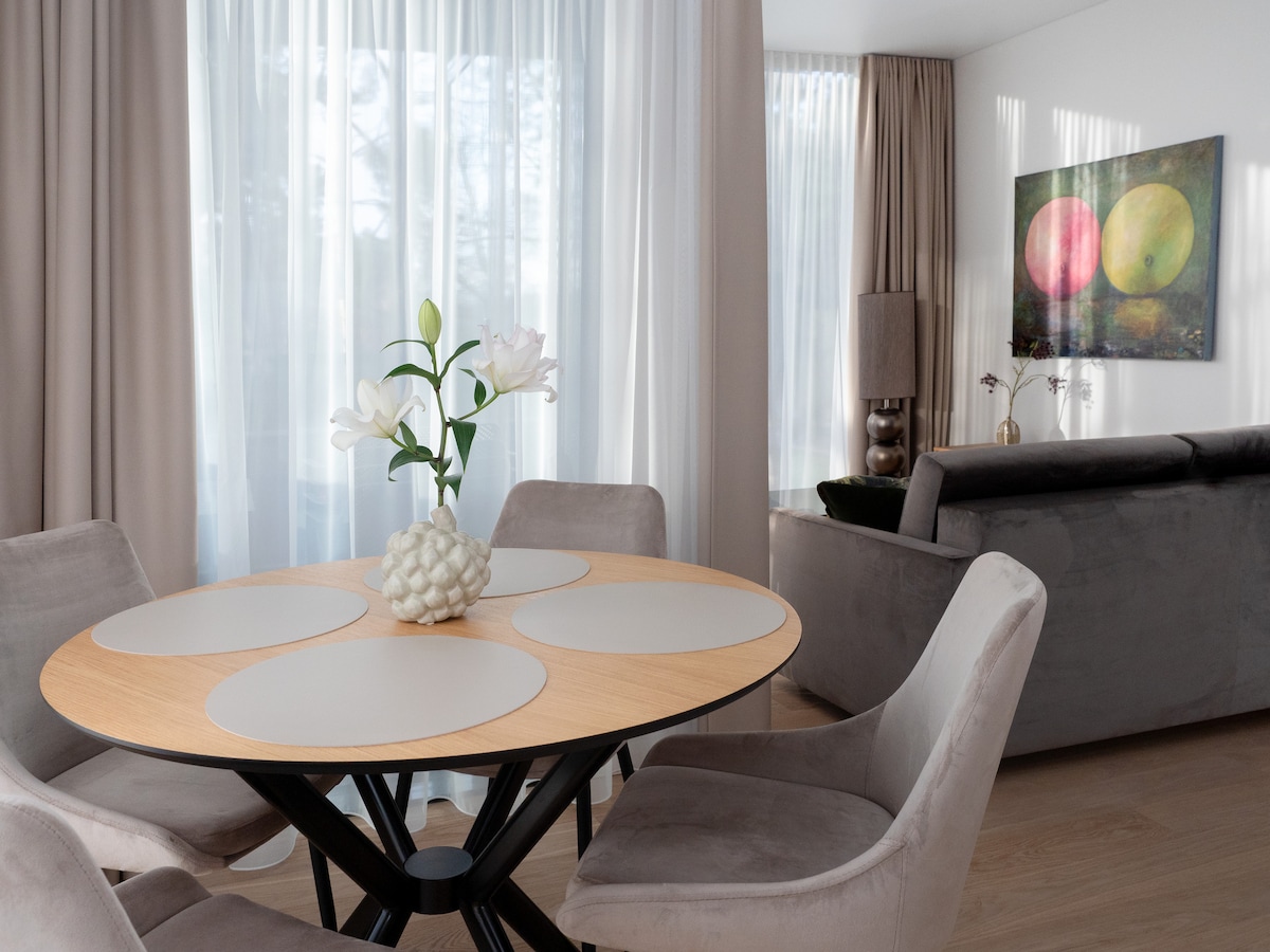 A dining area features a round wooden table with four placemats placed in front of light-colored chairs. A vase with flowers sits at the center of the table. Soft curtains allow natural light to brighten the space, while a colorful artwork is displayed on the wall.