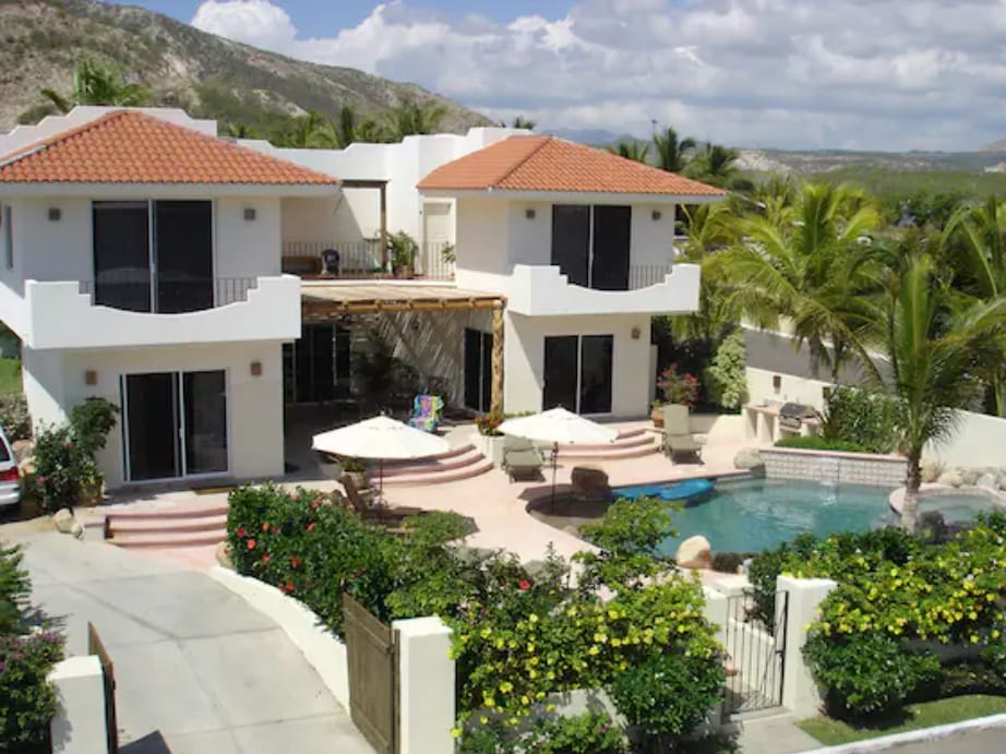 A two-story villa is pictured, featuring spacious balconies and large glass doors that provide views of the surrounding landscape. A private pool is set among lush greenery, with vibrant flowering plants framing the outdoor area. A patio umbrella offers shade near the pool.