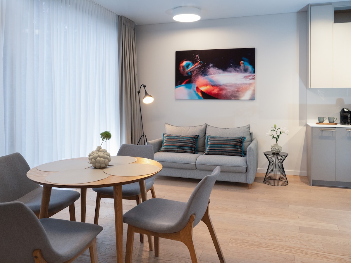 A dining area features a circular table surrounded by four grey chairs. A light grey sofa with decorative cushions is positioned against the wall. A piece of modern art hangs above, and a small plant adds a touch of greenery. Soft lighting and large windows allow natural light to fill the space.