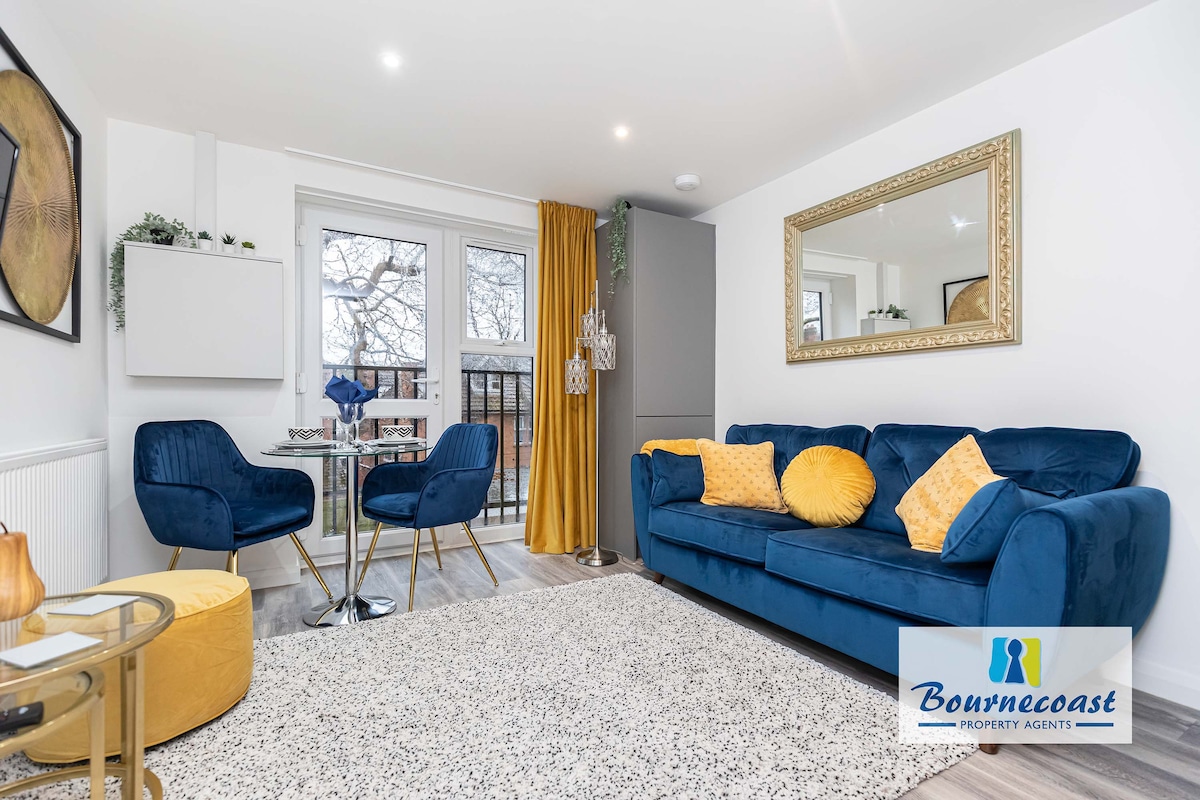 A modern lounge area is equipped with a blue sofa complemented by yellow cushions. A round dining table with two chairs is positioned beside floor-to-ceiling doors that open to a Juliette balcony. A decorative mirror and soft drapes enhance the ambiance.