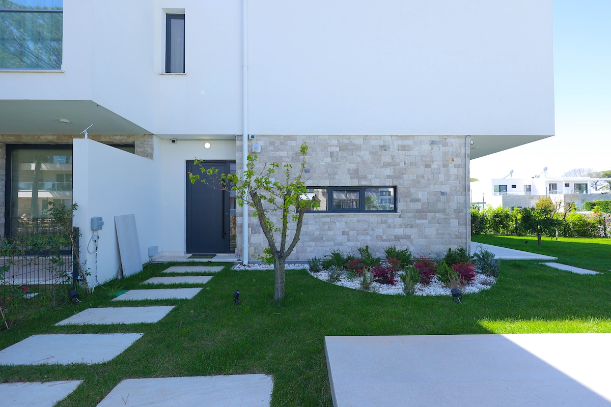 The exterior of a modern vacation rental features a landscaped garden with neatly arranged stone pathways leading to a dark entrance door. A small tree is planted in the yard, surrounded by vibrant plants and white stones.