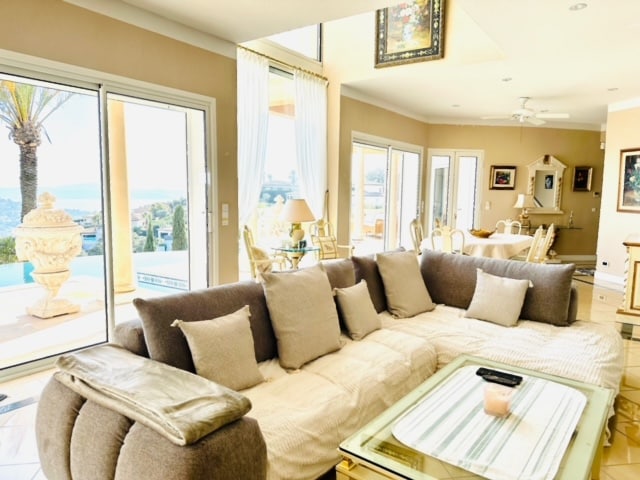 A spacious living area features a large, plush sectional sofa adorned with numerous cushions. Sunlight floods the space through expansive glass doors, revealing a view of the pool and landscape beyond. A glass coffee table sits in front, while a dining area is visible in the background.