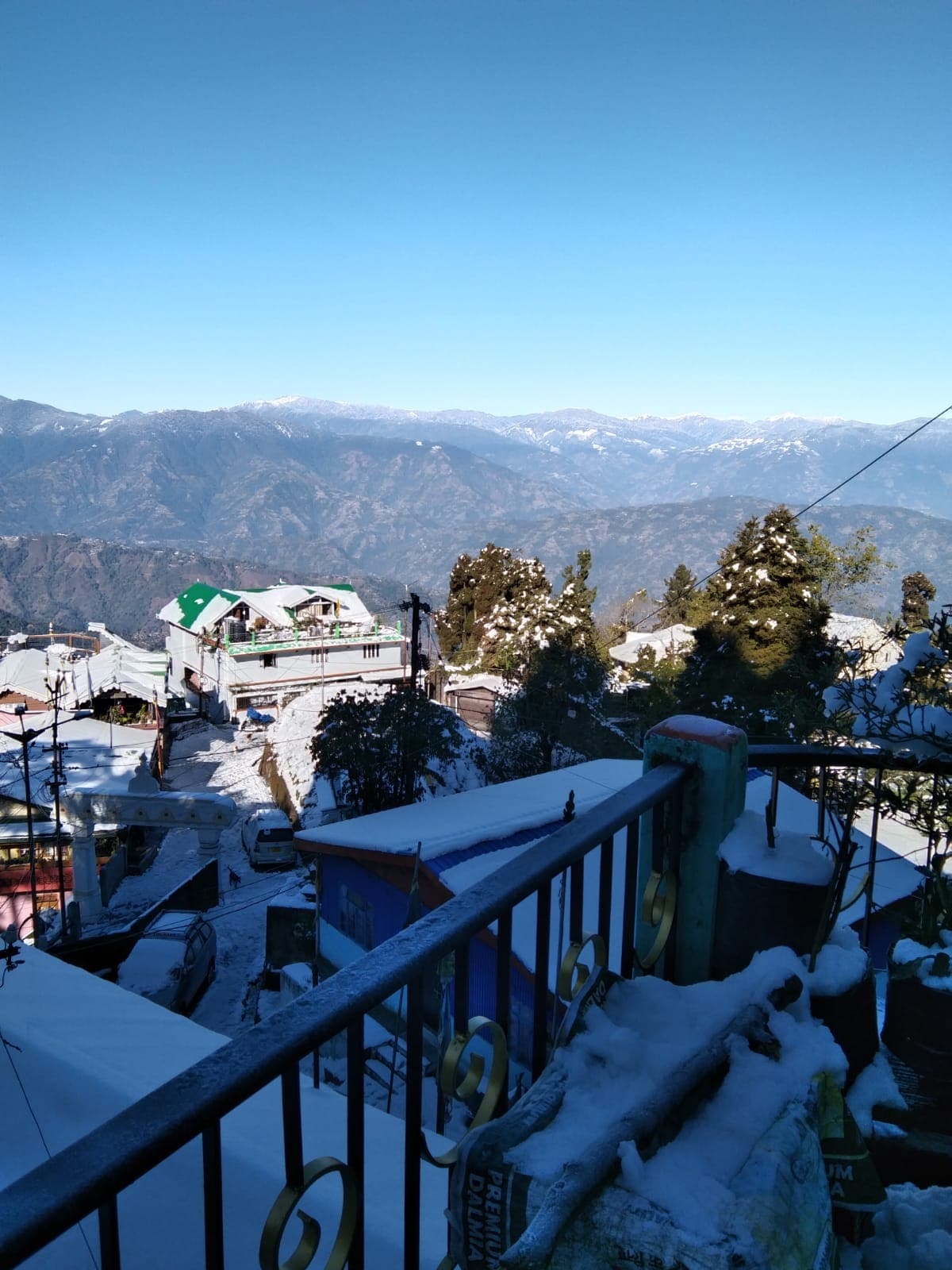 A panoramic view of snow-covered rooftops and pine trees is presented against a backdrop of majestic mountains. Clear blue skies provide a striking contrast to the white landscape, offering a serene, wintry atmosphere for relaxation.