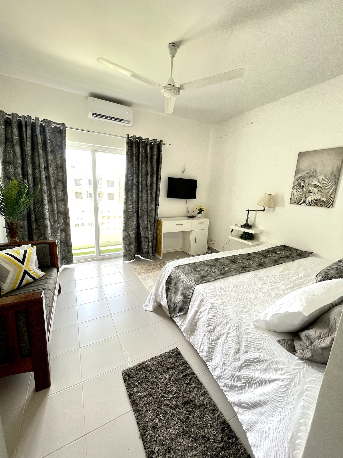 A bright studio features a spacious bed adorned with a textured white coverlet. A small seating area includes a wooden sofa with decorative cushions. Natural light enters through the glass doors, which lead to a patio, while a cozy rug lies beneath.