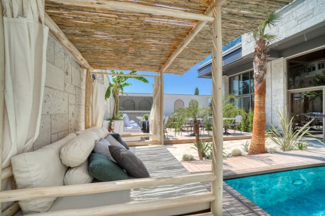 A shaded outdoor lounging area features a wooden structure with soft cushions, providing a comfortable space for relaxation. Tropical plants surround the area, alongside a sparkling pool that enhances the tranquil ambiance of the outdoors.
