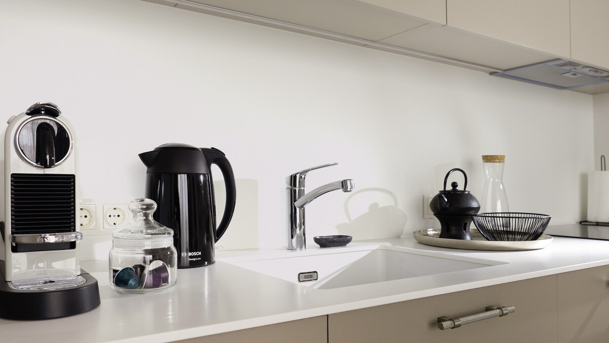 The kitchen area features a modern design, showcasing a countertop with an induction stove and sink. A Nespresso coffee machine, electric kettle, and stylish teapot are neatly arranged beside a dish rack. Additional dining items are displayed in a woven basket.