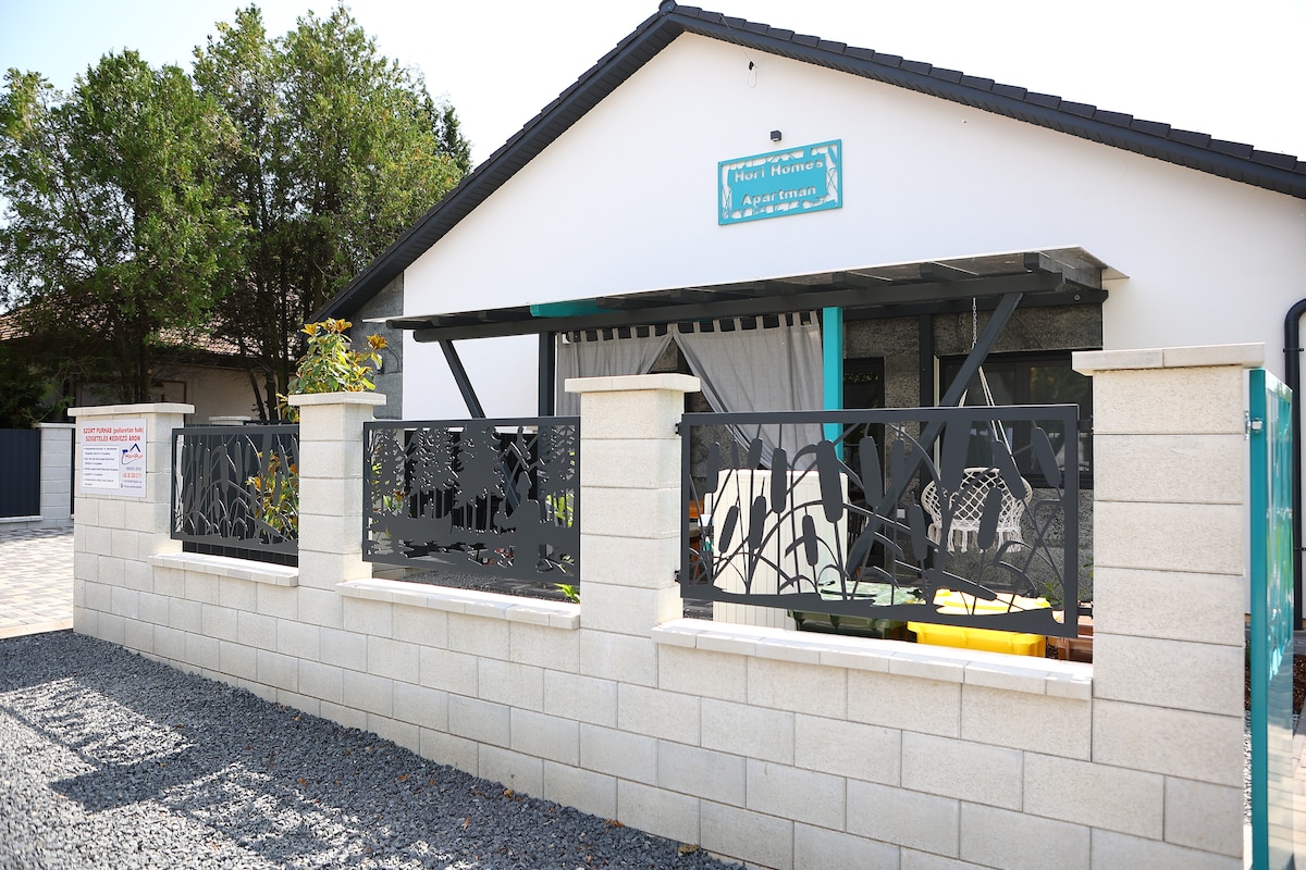 The exterior of a stylish accommodation is showcased, featuring a decorative stone wall with metal railings. A welcoming sign is displayed above the entrance, and the surrounding area is landscaped with greenery. A shaded porch area with seating is visible, inviting outdoor relaxation.