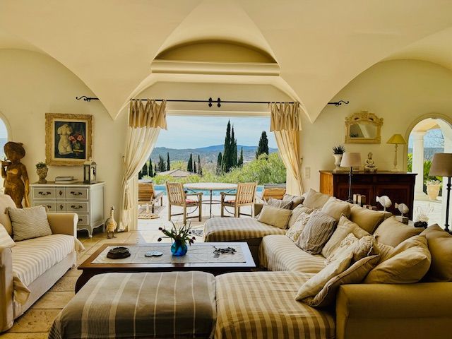 A spacious living area is highlighted by a large, comfortable sectional sofa adorned with patterned cushions. A wooden coffee table anchors the space, complemented by soft lighting and decorative accents. Arched windows frame a scenic outdoor view of lush greenery and distant hills.