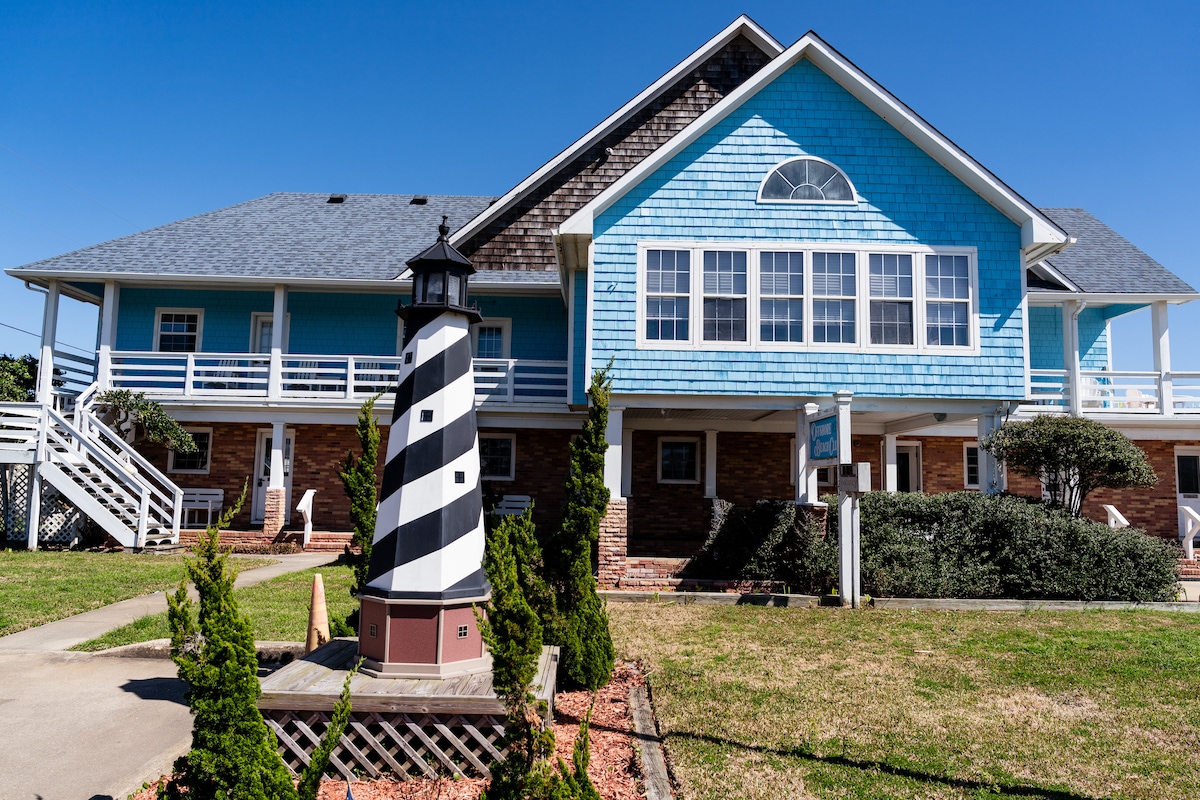 A charming condotel suite is visible with a blue façade and large windows, complemented by pristine landscaping. In the foreground, a classic lighthouse structure with black and white stripes adds a nautical touch to the scenery, enhancing the welcoming atmosphere of the property.