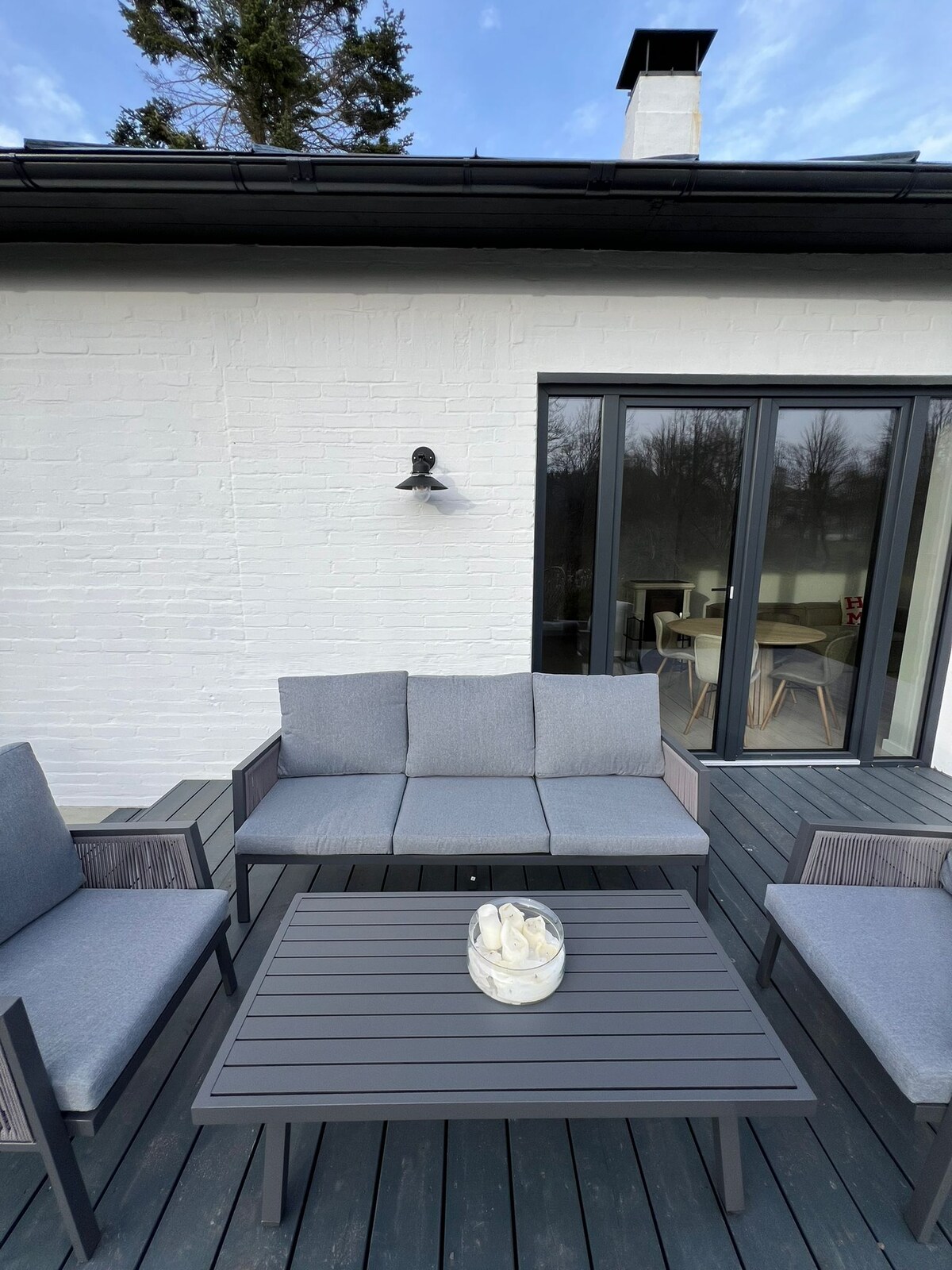 A patio space is furnished with a gray outdoor sofa and two matching chairs, arranged around a rectangular table. The table holds a decorative bowl, and soft natural light enhances the peaceful atmosphere. A backdrop of white wall and large glass doors is visible.